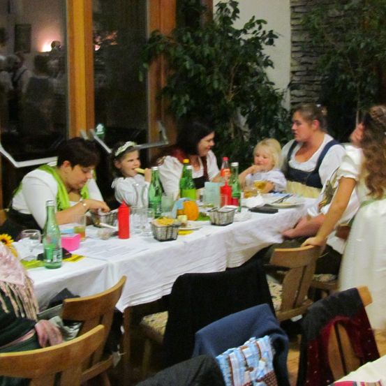 Mehrere Erwachsene und Kinder sitzen um einen Tisch herum und essen, mit verschiedenen Getränken und Speisen auf dem Tisch und Pflanzen im Hintergrund.