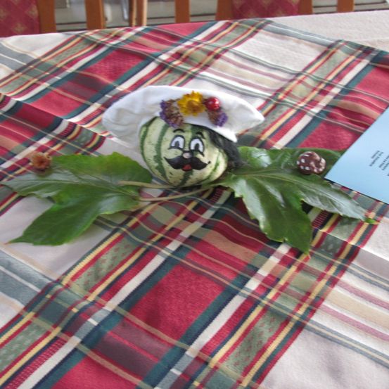 Eine Wassermelonenpuppe mit Kochmütze und Blumen sitzt auf einer karierten Tischdecke. Die Puppe hat grüne Blätter und kleine dekorative Pilze.