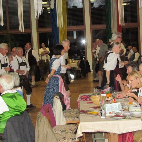 Eine Gruppe älterer Menschen versammelt sich in einem Raum, möglicherweise für eine Party. Einige stehen, andere sitzen. Sie tragen traditionelle Kleidung. Es gibt Tische mit Weingläsern, Tellern und Schüsseln. Der Raum hat Säulen und Fenster mit Vorhängen.