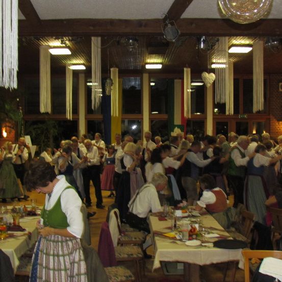 Eine große Gruppe von Menschen in traditioneller Kleidung versammelt sich in einem Saal. Einige tanzen, während andere klatschen und zuschauen. Tische mit Tellern, Gläsern und Blumen sind im Saal aufgestellt.