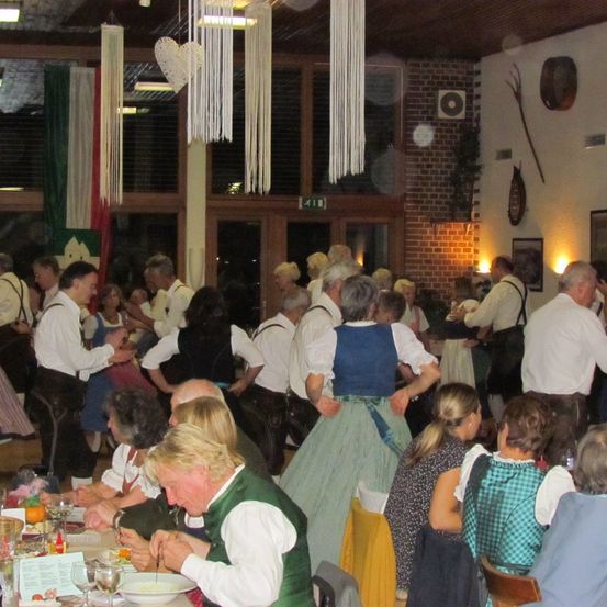 Eine Gruppe von Menschen in traditioneller deutscher Kleidung ist drinnen, sitzt an Tischen und steht in einem Raum. Der Raum hat Backsteinwände, dekorative Objekte und eine grüne Fahne.