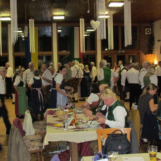 Eine Gruppe von Menschen versammelt sich in einem Speisesaal, einige stehen und andere sitzen um Tische herum. Der Raum ist mit verschiedenen hängenden Dekorationen geschmückt, und Flaggen zieren die Wände. Auf den Tischen befinden sich Gläser, Teller und Schüsseln, und ein Rucksack ist auf einem der Stühle abgestellt.