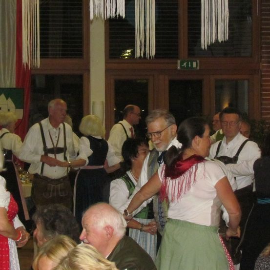 Eine Gruppe von Menschen ist in einem Raum versammelt. Einige tragen traditionelle Kleidung, wobei eine Frau tanzt. Der Raum ist mit weißen und roten Vorhängen dekoriert, und über der Tür befindet sich ein grünes Schild.