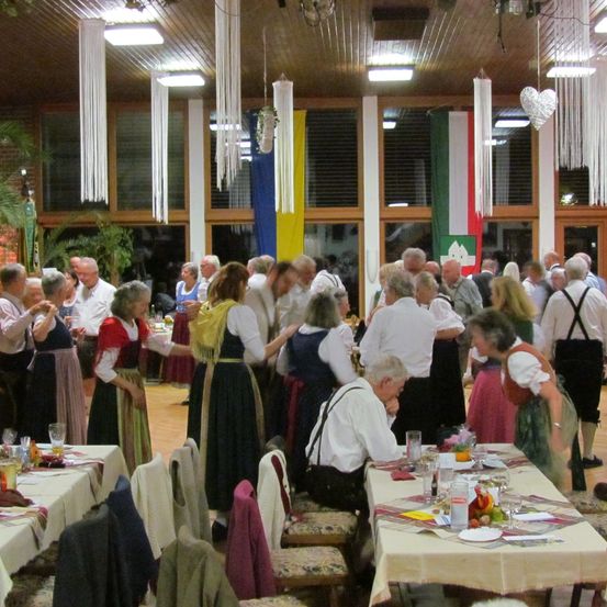 Ein Raum mit vielen Menschen in traditioneller Kleidung. Tische sind mit Tellern, Gläsern und Servietten gedeckt. Einige Personen stehen, andere sitzen. Die Decke hat hängende Lampen und Flaggen.