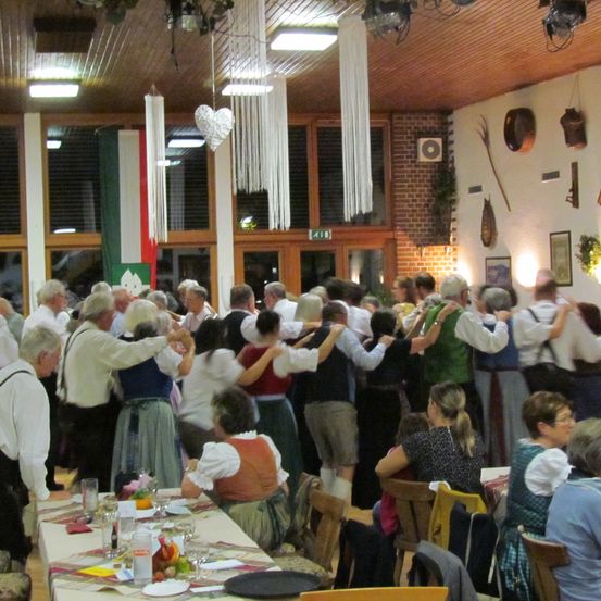 Eine Gruppe von Menschen in traditioneller bayerischer Kleidung versammelt sich in einem Saal, wahrscheinlich für eine kulturelle Veranstaltung. Sie tanzen paarweise, während andere an Tischen sitzen. Der Raum hat eine rustikale Einrichtung mit Holzbalken und dekorativen Gegenständen an den Wänden.