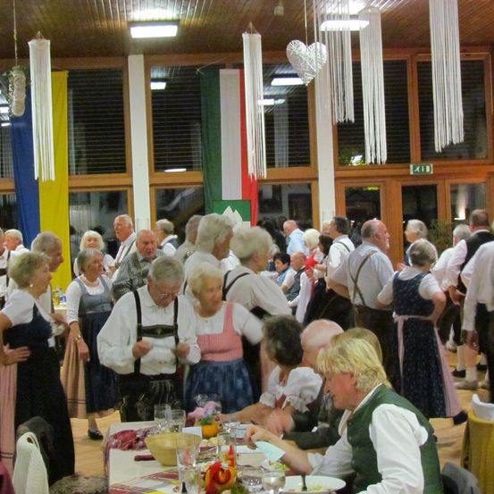 Eine große Gruppe älterer Menschen in traditioneller Kleidung versammelt sich in einem Saal, unterhält sich und isst. Der Raum ist mit verschiedenen Dekorationen wie Flaggen und hängenden Quasten geschmückt. Die Tische sind mit Tellern, Weingläsern und Speisen gedeckt.