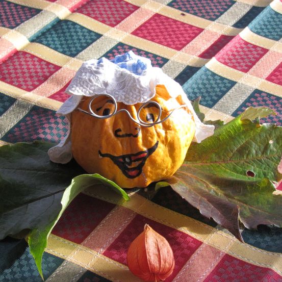 Ein Kürbis mit Gesicht, der eine Haube und eine Brille trägt, liegt auf einer bunten Tischdecke mit Herbstblättern.