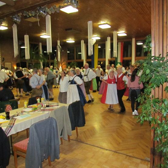Ein Saal voller tanzender Menschen, einige in traditionellen Kleidern. Ein Tisch mit Getränken und Essen. Lichter und Pflanzen schmücken den Raum.