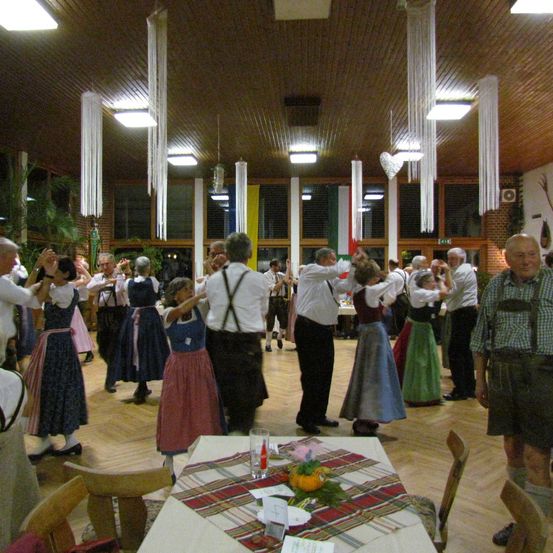 Eine Gruppe älterer Erwachsener in traditioneller Kleidung tanzt in einem Raum mit Holzböden, umgeben von Tischen und Stühlen, mit dekorativen Elementen, die von der Decke hängen.