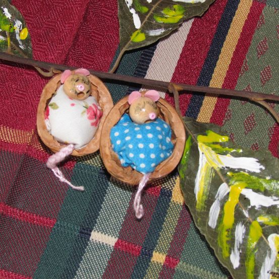 Zwei Mausspielzeuge sind in Walnussschalen platziert und an einem Zweig aufgehängt. Sie liegen auf einem bunten, gemusterten Tuch.