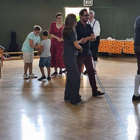 Eine Gruppe von Menschen tanzt in einer Halle. Eine Frau in einem roten Kleid und ein Mann in einem schwarzen Shirt führen den Tanz an. Ein Tisch mit Sonnenblumen steht hinter ihnen.