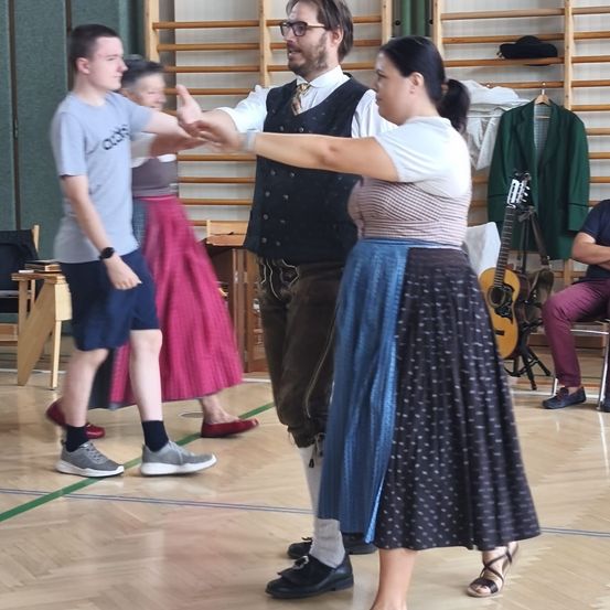 Eine Gruppe von Menschen tanzt in einem Raum. Ein Mann und eine Frau führen den Tanz an.