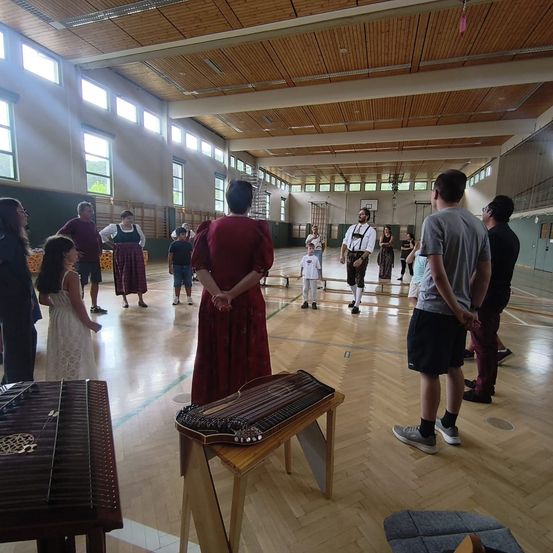 Eine Gruppe von Menschen in einer Turnhalle, einige stehen und andere sitzen. Eine Frau in einem roten Kleid steht vor einem Holztisch mit einem Musikinstrument. Ein Kind steht vor einer Bank. Die Turnhalle hat Holzböden und weiße Wände.