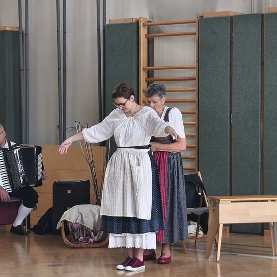Zwei Frauen in traditioneller Kleidung tanzen, während ein Mann Akkordeon spielt in einem Raum mit Holzmöbeln und grünen Trennwänden.