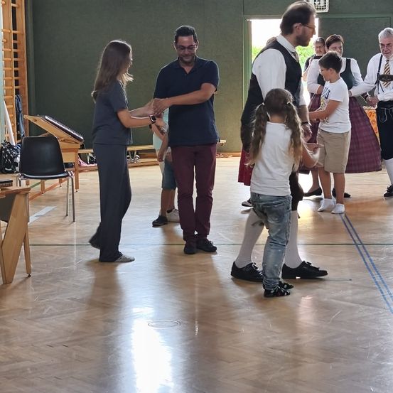 Eine Gruppe von Menschen in einem Raum, einige tanzen, während andere stehen und zusehen. Ein Mann und eine Frau schütteln sich die Hände.