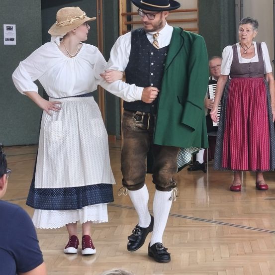 Ein Mann und eine Frau in traditioneller bayerischer Kleidung tanzen zusammen auf einem Holzboden. Eine ältere Frau in ähnlicher Kleidung steht in der Nähe, und ein Mann spielt Akkordeon.