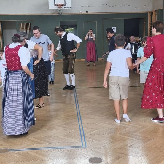 Eine Gruppe von Menschen in traditioneller Kleidung tanzt in einer Turnhalle mit einem Basketballkorb im Hintergrund.