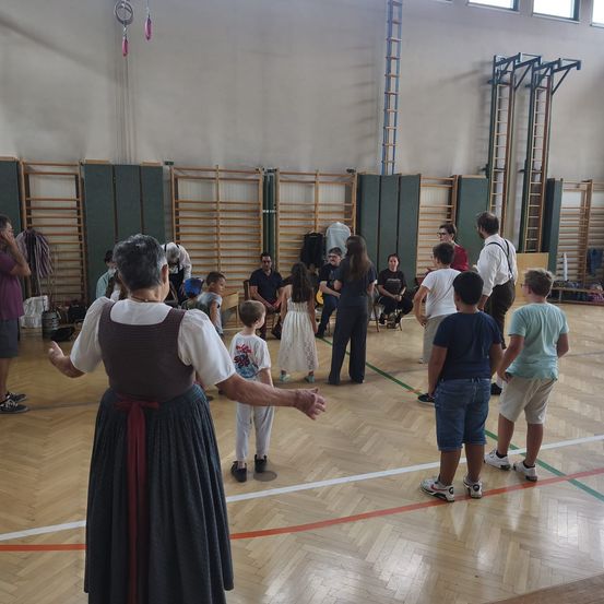Eine Gruppe von Menschen, einschließlich Kindern, versammelt sich in einer Turnhalle. Eine ältere Frau in traditioneller Kleidung führt einen Tanz an.