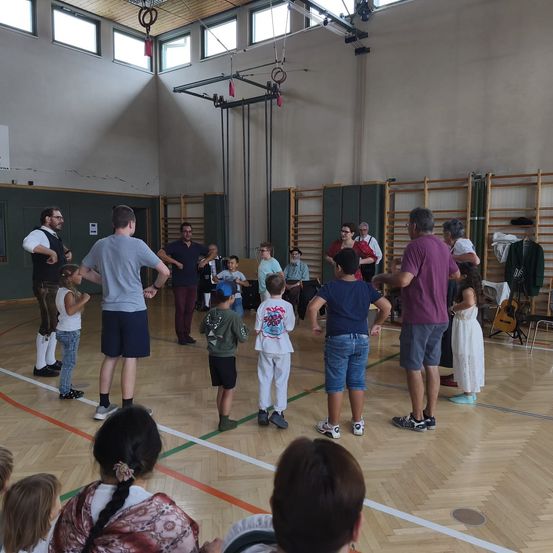 Eine Gruppe von Erwachsenen und Kindern steht in einem Kreis in einer Turnhalle, möglicherweise üben sie einen Tanz oder eine Aufführung. Die Erwachsenen stehen vor den Kindern, und einige halten Musikinstrumente. Die Kinder tragen Freizeitkleidung. Die Turnhalle hat Holzböden, eine grüne Wand und eine weiße Decke.