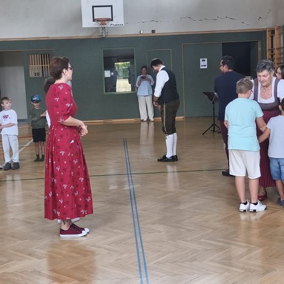 Eine Gruppe von Menschen versammelt sich in einer Turnhalle. Eine Frau in einem roten Blumenkleid steht in der Mitte, während andere, einschließlich Kinder, um sie herum stehen. Ein Mann in einem Weste und Lederschuhen steht in der Mitte des Raumes. Ein Basketballkorb ist an der Wand montiert.