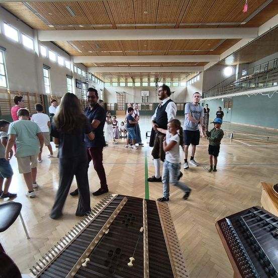 Eine Gruppe von Menschen, einschließlich Kindern, versammelt sich in einer Turnhalle. Einige stehen, andere gehen, und es gibt Musikinstrumente auf dem Boden.