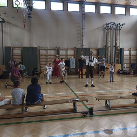 Eine Gruppe von Menschen versammelt sich in einer Turnhalle. Ein Mann in traditioneller bayerischer Kleidung steht in der Mitte und gestikuliert mit den Händen. Kinder und Erwachsene sind verstreut, einige sitzen auf Bänken, während andere stehen oder auf dem Boden sitzen. Verschiedene Turngeräte sind im Hintergrund zu sehen.