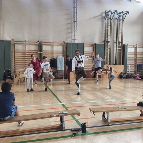 Eine Gruppe von Menschen übt eine Tanzroutine in einer Turnhalle. Ein Mann in einem weißen Hemd und Hosenträgern springt in die Luft.