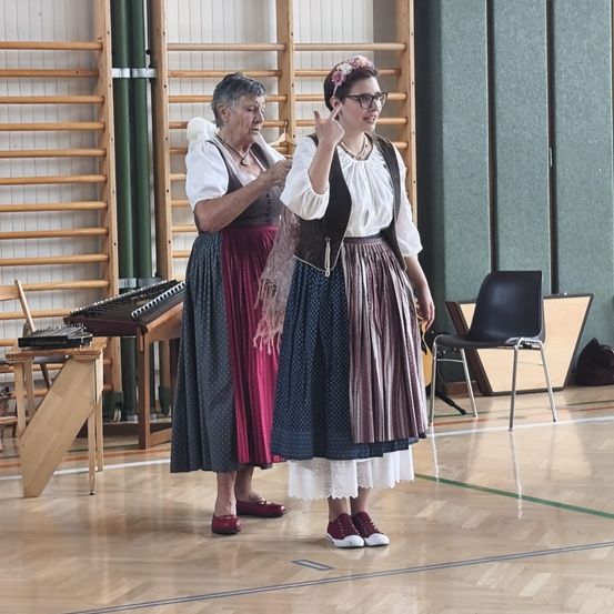 Zwei Frauen in traditioneller Kleidung stehen in einer Turnhalle, eine zeigt mit der Hand. Hinter ihnen steht ein Holztisch mit Musikinstrumenten.