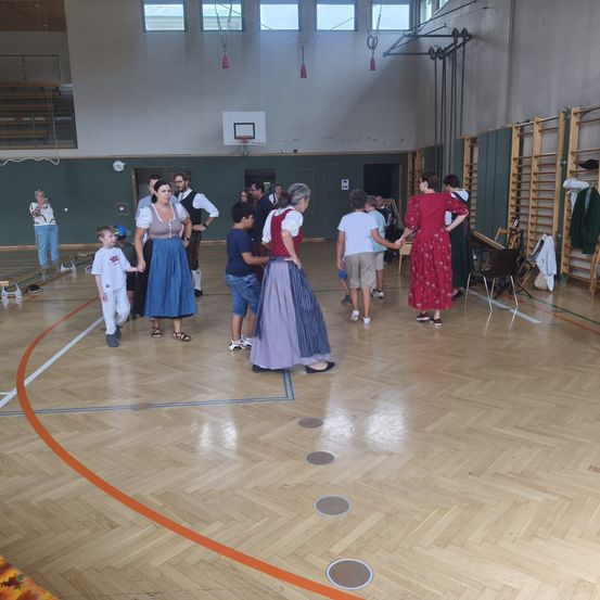 Eine Gruppe von Menschen tanzt in einer Turnhalle, einige in traditioneller Kleidung. Ein Basketballkorb ist an der Wand montiert.