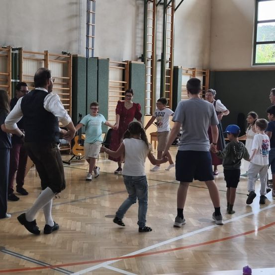 Eine Gruppe von Menschen, einschließlich Erwachsener und Kindern, tanzt in einer Turnhalle mit Holzböden. Einige halten sich an den Händen, und einige tragen Hüte. Musikinstrumente sind an der Seite platziert.