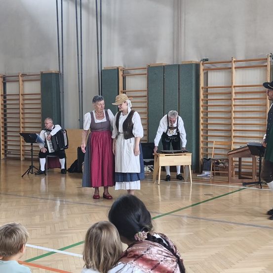 Eine Gruppe von Menschen in traditioneller Kleidung führt auf einer Bühne mit Musikinstrumenten auf. Ein Mann spielt Akkordeon, während eine Frau neben einer anderen steht. Dahinter befindet sich eine Holzstruktur mit einem Klavier obenauf.