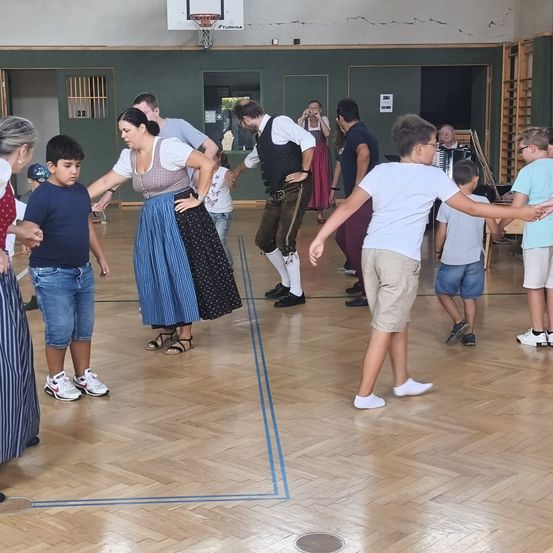 Eine Gruppe von Menschen, einschließlich Kinder, tanzen in einer Turnhalle. Einige tragen traditionelle Kleidung. Eine Person spielt Akkordeon.