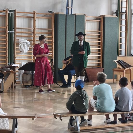 Eine Frau in einem roten Kleid und ein Mann in einem grünen Anzug stehen vor einer Gruppe von Kindern, die auf einer Bank sitzen. Dahinter sitzt ein Mann auf einem Stuhl und spielt Gitarre.