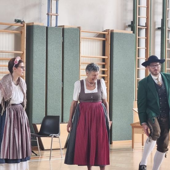 Drei Personen in traditioneller Kleidung stehen in einem Raum mit grünen Wänden und Holztrennwänden. Eine Frau mit einer Blume im Haar und einer Brille schaut nach links. Ein Mann in einem grünen Anzug und Hut steht rechts. Eine Frau in einem rot-weißen Kleid steht in der Mitte.