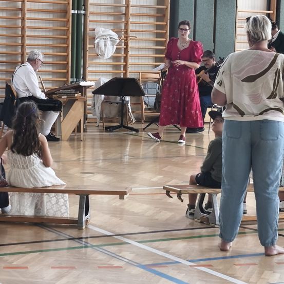Eine Gruppe von Menschen versammelt sich in einem Raum. Eine Frau in einem roten Kleid steht und spricht, während ein Mann in einem weißen Hemd sitzt und ein Musikinstrument spielt. Ein Mädchen in einem weißen Kleid sitzt auf einer Bank. Eine andere Frau steht und schaut die Frau im roten Kleid an.