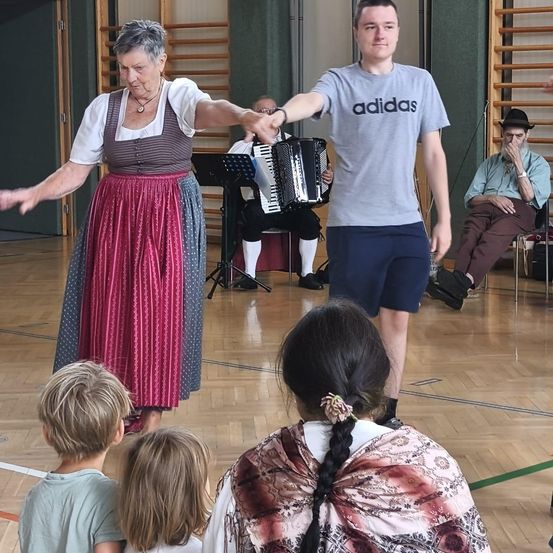 Eine Gruppe von Menschen versammelt sich in einem Raum. Eine ältere Frau in einem traditionellen Kleid tanzt mit einem Mann in einem Adidas-Shirt. Ein Mann spielt Akkordeon, während er sitzt. Ein Mann sitzt auf einem Stuhl mit der Hand im Gesicht.