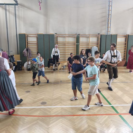 Eine Gruppe von Kindern und Erwachsenen tanzt in einer Turnhalle mit Holzböden und weißen Wänden. Einige Erwachsene stehen und beobachten.