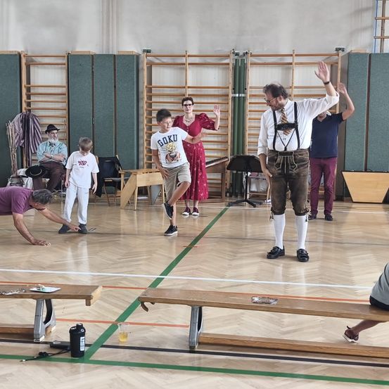 Eine Gruppe von Menschen in einer Turnhalle, ein Mann in traditioneller bayerischer Kleidung winkt, während andere in verschiedenen Posen stehen. Holzbänke und eine grüne Linie auf dem Boden sind sichtbar.