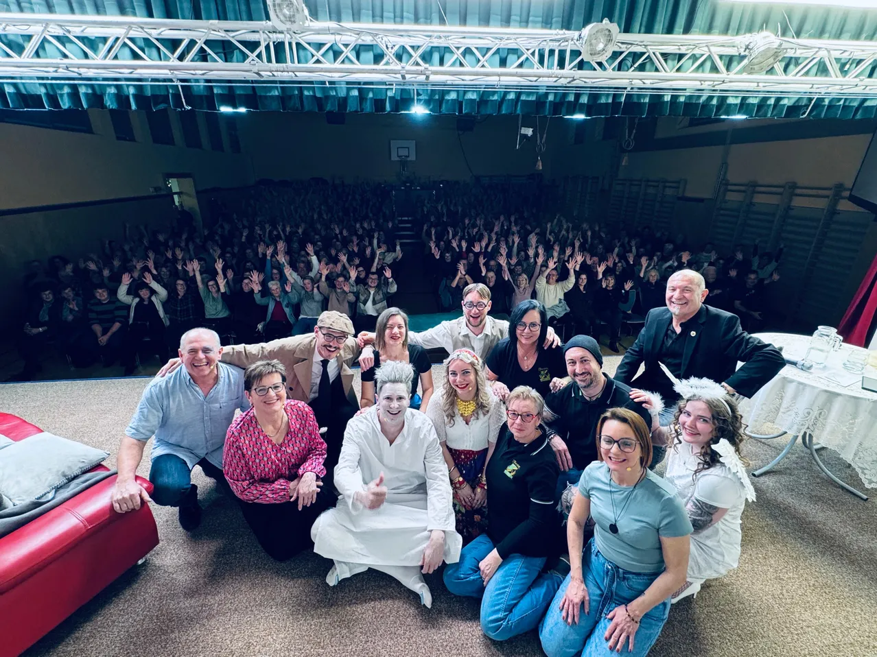 Eine Gruppe von Menschen posiert für ein Foto in einem großen Saal mit einem Publikum im Hintergrund. Einige stehen, andere sitzen, mit einer Person in weißer Kleidung in der Mitte, die lächelt.
