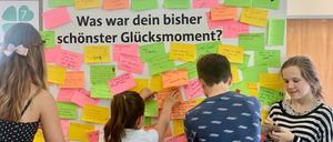 Drei Menschen stehen vor einer Wand mit bunten Haftnotizen. Ein Mädchen und ein Junge schauen sich die Notizen an, während die dritte Person ein Buch betrachtet. Die Wand trägt den Text 'Was war dein bisher schonster Glücksmoment?'.