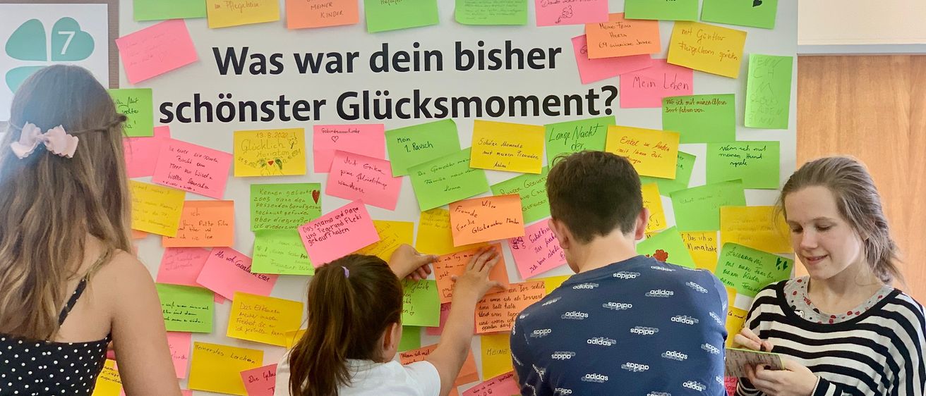 Drei Menschen stehen vor einer Wand mit bunten Haftnotizen. Ein Mädchen und ein Junge schauen sich die Notizen an, während die dritte Person ein Buch betrachtet. Die Wand trägt den Text 'Was war dein bisher schonster Glücksmoment?'.