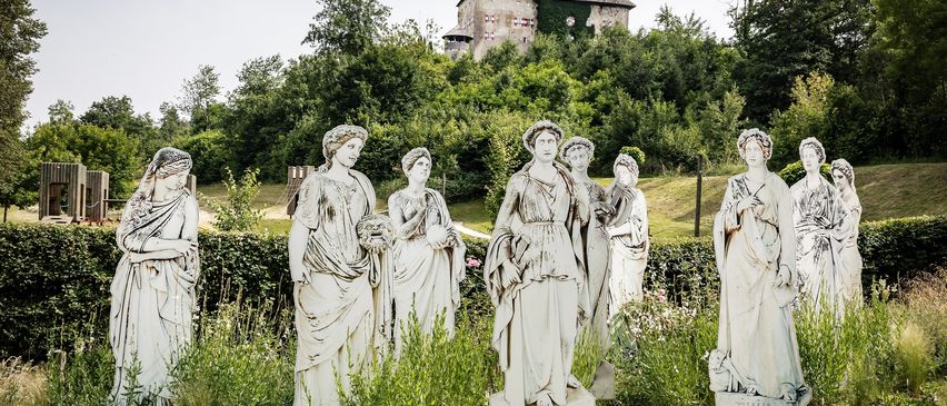 Eine Gruppe weißer Statuen von Frauen steht in einem Garten, mit einem Haus und Bäumen im Hintergrund.