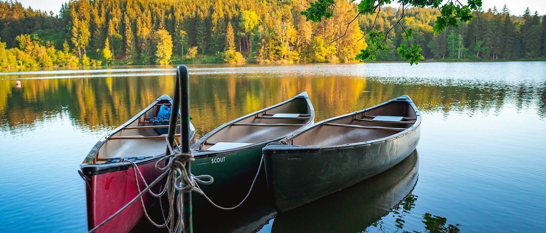 Drei Kanus sind an einem Steg auf einem ruhigen See mit Wald im Hintergrund, unter einem klaren Himmel, festgemacht.