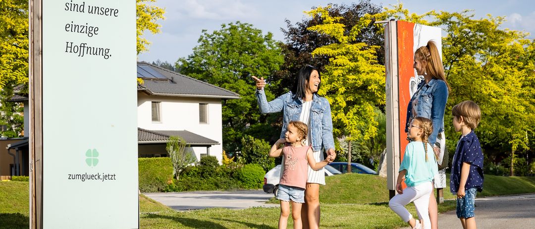 Eine Familie mit zwei Kindern steht vor einem Haus mit Solarpanelen. Sie lächeln und scheinen für ein Foto zu posieren. Dahinter befindet sich eine Rasenfläche mit Bäumen.