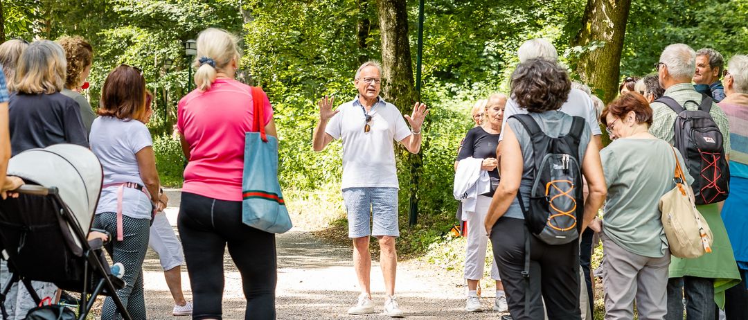 Eine Gruppe von Menschen steht auf einem Kiesweg in einem Park, ein Mann spricht mit erhobenen Händen. Einige tragen Rucksäcke und manche tragen Turnschuhe. Bäume umgeben das Gebiet.