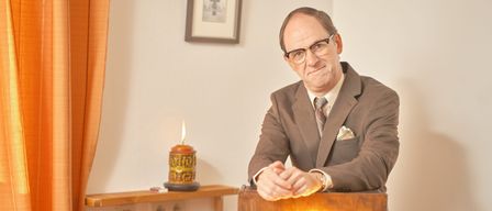 Ein Mann steht lächelnd vor einem beleuchteten Holzschild. Dahinter befindet sich ein Regal mit einer brennenden Kerze und einem Bilderrahmen an der Wand.