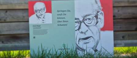 Ein Poster mit einer Zeichnung eines Mannes und Text in Deutsch steht auf einer Grasfläche vor einer Holzwand.