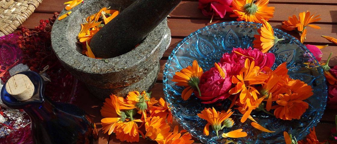 Ein Mörser und Stößel mit zerdrückten Blumen, auf einer Holzoberfläche platziert. Daneben eine blaue Schüssel mit zusätzlichen Blumen.