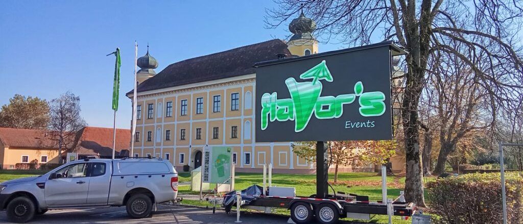Ein Anhänger mit einem Schild für Nayors Events steht vor einem großen, gelb gestrichenen Gebäude. Ein Pickup-Truck ist in der Nähe geparkt.