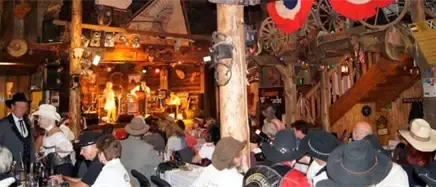 Eine rustikale Indoor-Veranstaltung mit Bühne, Leuten in Hüten und Anzügen, Holzdekor und Flaggen. Es gibt Tische und Stühle, eine Treppe und eine offene Bar.